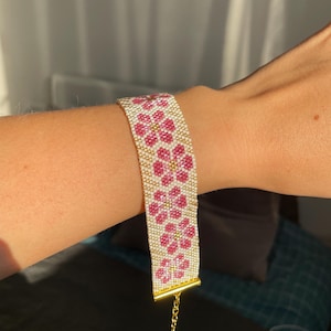 May include: A wide beaded bracelet featuring a repeating floral pattern with pink petals and gold centers against a cream-colored background. The bracelet is finished with a gold-toned metal bar clasp and a delicate gold-toned chain extender.