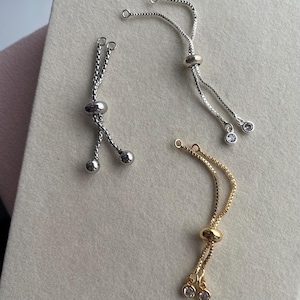May include: Three adjustable jewellery sliders with box chains. One silver-tone slider features plain round end beads, while the other silver-tone and the gold-tone sliders each feature two small clear round stones at the ends of the chains.