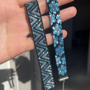 May include: Two beaded bracelets with blue, black, and silver patterns. One bracelet has a zigzag design, the other a floral pattern. Both have a black backing and silver clasps. The bracelets are likely around 15-20 cm in length.