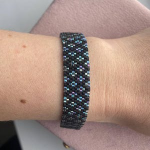 May include: A black beaded bracelet with iridescent highlights. The bracelet is made of small, tightly woven beads, creating a textured surface. The iridescent beads reflect a range of colours, including blue, purple, and green. The bracelet is approximately 2.5 cm wide.