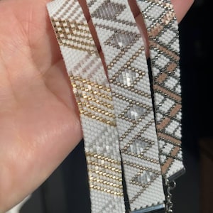 Può includere: Quattro braccialetti di perline bianche, dorate, argentate e beige. I braccialetti hanno motivi geometrici e sono collegati con una catena d'argento. I braccialetti sono fatti di piccole perline.