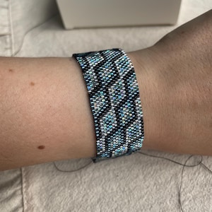 Può includere: Un braccialetto di perline con un motivo geometrico nei toni del verde acqua, argento e nero è indossato su un polso. Il braccialetto è fatto di piccole perline distanziate in modo uniforme. Una scatola bianca è sullo sfondo.