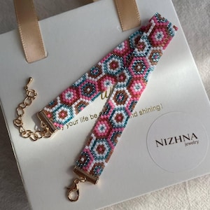Puede incluir: Una pulsera de cuentas con un patrón geométrico en rosa, turquesa, marrón y blanco. La pulsera tiene extremos de metal dorado y una cadena. Está sobre una caja de regalo blanca con una cinta dorada y el texto "your life be... and shining!" y la marca "NIZHNA jewelry".