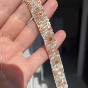 May include: A beaded bracelet in a flat, woven style. The bracelet features a pattern of light pink, white, and gold beads. The design includes small, gold-colored floral accents. The bracelet is held in a hand.