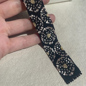 May include: A flat, rectangular beaded bracelet featuring a black background with a repeating floral-inspired pattern in muted beige and metallic gold seed beads. The design is composed of small, uniform beads arranged in a grid-like structure.