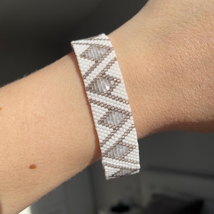 May include: White beaded bracelet with a geometric pattern in shades of gray and gold. The bracelet is made of small, evenly spaced beads, creating a textured surface. The design features a repeating diamond pattern.