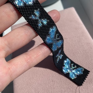 May include: A woven bead bracelet featuring a black background with a repeating pattern of iridescent blue butterflies. The bracelet is constructed from small, uniform beads arranged in a grid-like structure, creating a flat, flexible band.