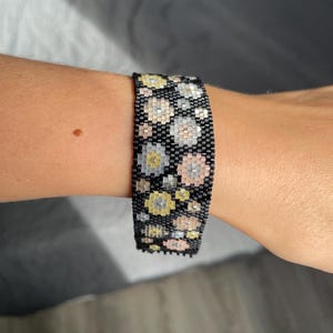 May include: A wide cuff bracelet crafted from small black seed beads featuring a repeating pattern of circular floral motifs in shades of pale yellow, soft pink, and metallic silver, arranged in a dense, grid-like woven structure.