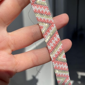 Puede incluir: Una pulsera de cuentas tejidas con un patrón de chevrón repetitivo en tonos rosa suave, amarillo pálido y blanco, acentuado con líneas finas de cuentas plateadas metálicas. La pulsera está construida con pequeñas cuentas cuadradas uniformes.