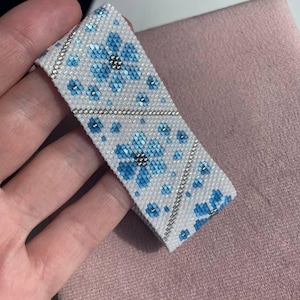Könnte beinhalten: Ein weißes Perlenarmband mit einem Blumenmuster in Hell- und Dunkelblau. Das Armband hat ein sich wiederholendes Design aus Blumen und kleinen blauen Perlen, wobei silberfarbene Perlen die Ränder umranden.