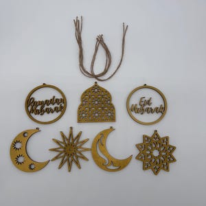 May include: Wooden Ramadan and Eid Mubarak ornaments in various shapes, including crescent moons, stars, and circular frames. The ornaments are light brown with intricate cut-out designs and include twine for hanging.