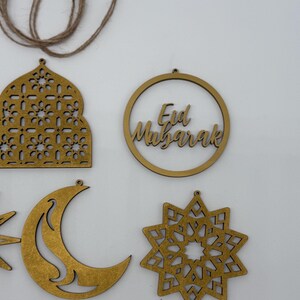 May include: Gold-colored wooden Eid Mubarak decorations. The image shows a crescent moon, a star, a mosque-shaped ornament, and a circular ornament with the text "Eid Mubarak". A length of twine is visible at the top.