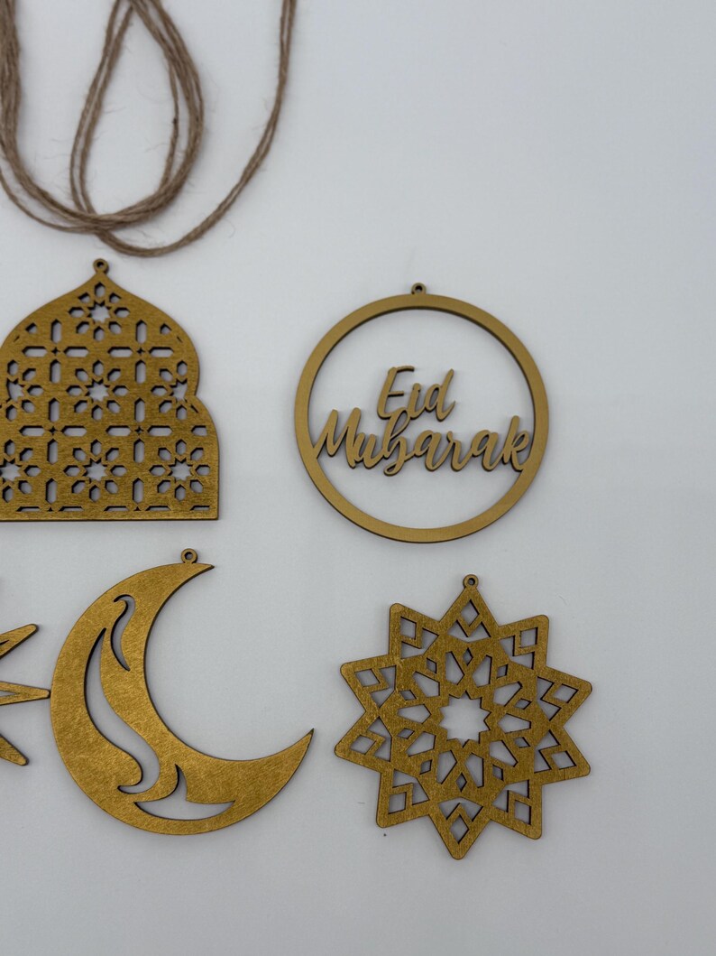 May include: Gold-colored wooden Eid Mubarak decorations. The image shows a crescent moon, a star, a mosque-shaped ornament, and a circular ornament with the text "Eid Mubarak". A length of twine is visible at the top.