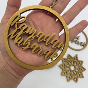 May include: Gold-colored wooden Ramadan Mubarak ornaments. The main ornament is a circular frame with the words "Ramadan Mubarak" in elegant script. Additional ornaments include a star-shaped design and a smaller circular ornament.