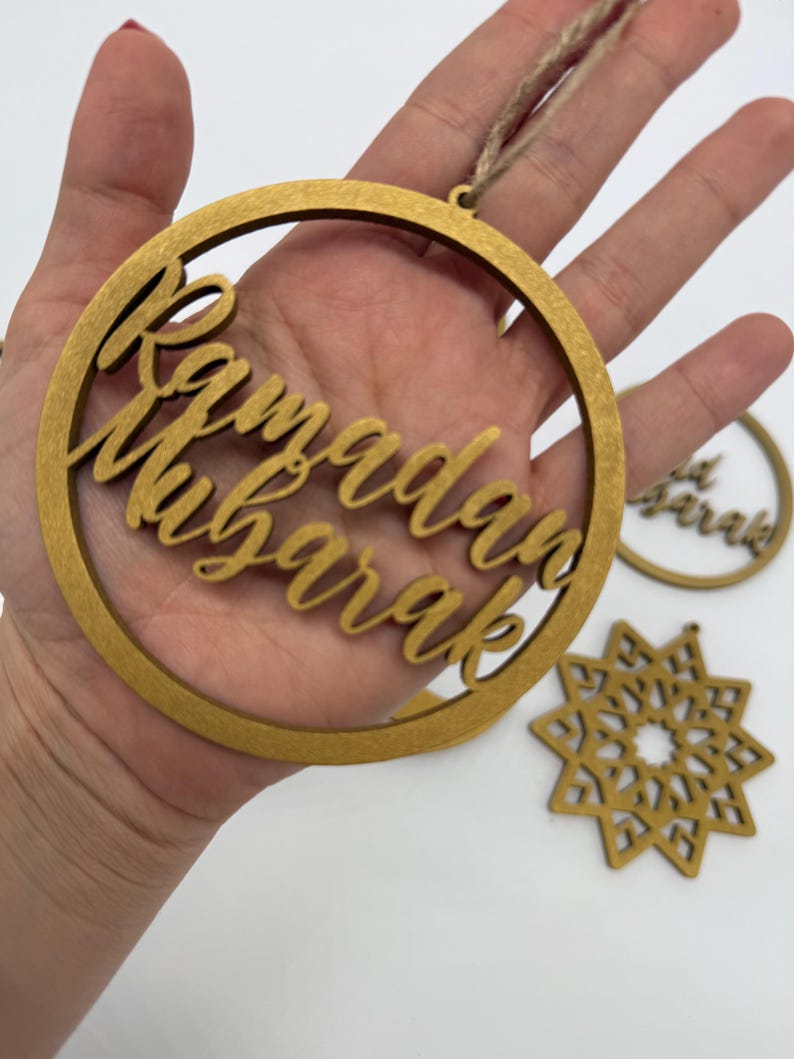 May include: Gold-colored wooden Ramadan Mubarak ornaments. The main ornament is a circular frame with the words "Ramadan Mubarak" in elegant script. Additional ornaments include a star-shaped design and a smaller circular ornament.