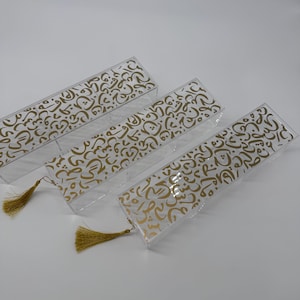 May include: Clear rectangular boxes with gold calligraphy patterns. Each box has a gold tassel attached. The boxes are arranged on a white surface. The calligraphy is in a repeating pattern.