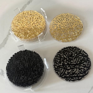 May include: A set of circular coasters featuring intricate Arabic calligraphy designs. Two coasters are finished in a metallic gold, and two are finished in a matte black. The coasters are displayed with a clear acrylic holder for storage.