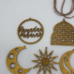 May include: Wooden Ramadan Mubarak decorations in various shapes, including a crescent moon, star, and a circular frame with the words "Ramadan Mubarak." The decorations are a light brown color and are designed to be hung.