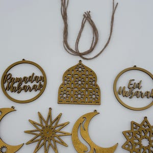 May include: A collection of wooden Ramadan and Eid Mubarak ornaments in various shapes, including crescent moons, stars, and circular frames with text. The ornaments are a light brown color and are connected to twine for hanging.