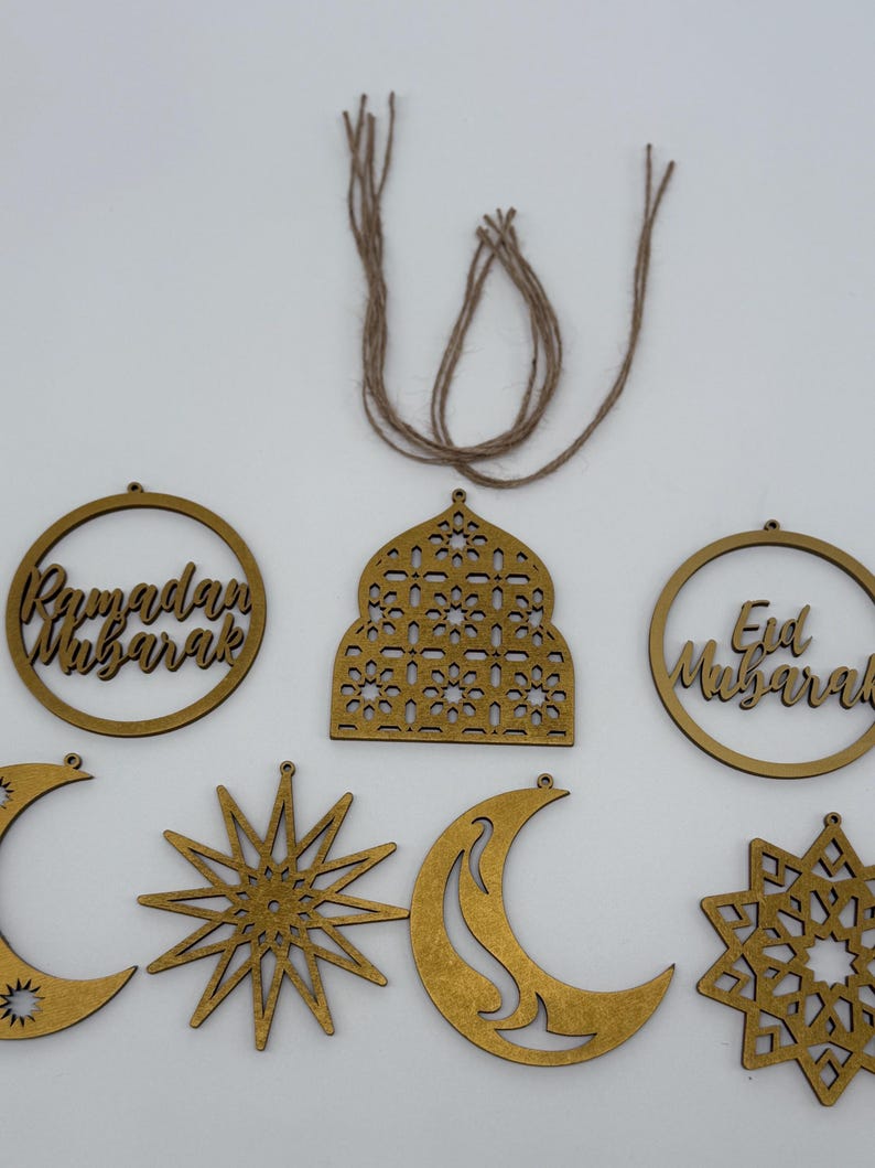 May include: A collection of wooden Ramadan and Eid Mubarak ornaments in various shapes, including crescent moons, stars, and circular frames with text. The ornaments are a light brown color and are connected to twine for hanging.