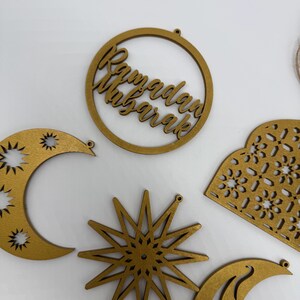 May include: Wooden Ramadan Mubarak ornaments in various shapes, including a crescent moon, star, and a circular design with the text "Ramadan Mubarak". The ornaments are gold-colored and have small holes for hanging.