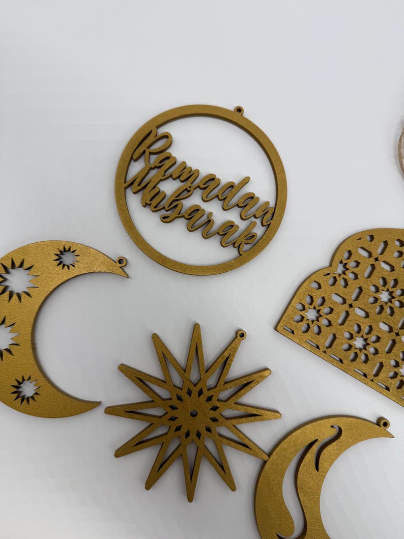 May include: Wooden Ramadan Mubarak ornaments in various shapes, including a crescent moon, star, and a circular design with the text "Ramadan Mubarak". The ornaments are gold-colored and have small holes for hanging.