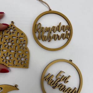 May include: Wooden Ramadan and Eid Mubarak ornaments. The gold-colored ornaments include a circular design with the words "Ramadan Mubarak" and "Eid Mubarak" in a script font. Other shapes include a mosque-shaped ornament and a crescent moon.