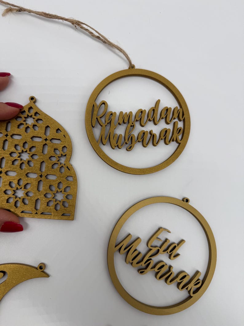 May include: Wooden Ramadan and Eid Mubarak ornaments. The gold-colored ornaments include a circular design with the words "Ramadan Mubarak" and "Eid Mubarak" in a script font. Other shapes include a mosque-shaped ornament and a crescent moon.