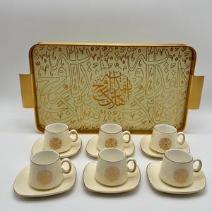 Puede incluir: Un juego de café color crema con seis tazas y platillos, y una bandeja a juego. La bandeja y las tazas presentan caligrafía árabe dorada. Las tazas y los platillos tienen un diseño circular dorado. La bandeja tiene asas doradas.
