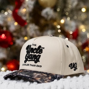 May include: A beige baseball cap with a camouflage brim. The cap features the text "The Uncle Gang Cooler Than Dad" in black script. The initials "UG" are on the side.