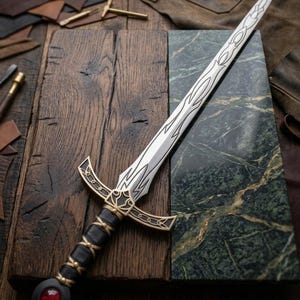 May include: A decorative sword with a silver blade featuring an etched design. The hilt is black with gold accents and a red gem at the pommel. The sword rests on a wooden surface.