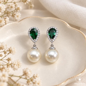 May include: A pair of drop earrings featuring a pear-shaped emerald green gemstone surrounded by a halo of small clear stones, suspended above a round white pearl. The setting is silver-toned metal, creating a classic and elegant jewelry piece.