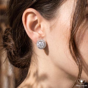 May include: A close-up of a sparkling, round stud earring with a halo of smaller stones. The earring is set in a silver-coloured metal and is worn on an ear. The jewelry is from Alice Delicate Jewelry Bar.