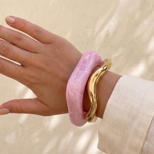 Può includere: Un set di due bracciali indossati al polso. Un bracciale è un rigido spesso in resina traslucida rosa chiaro, dalla forma liscia, arrotondata e irregolare. Il secondo bracciale è un rigido in metallo dorato lucido con un design ondulato.