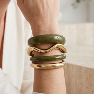 Può includere: Una pila di quattro braccialetti indossati al polso. Il set comprende due bracciali rigidi in resina liscia color verde oliva, un bracciale rigido in metallo dorato lucido dalla forma ondulata e un bracciale rigido in metallo dorato liscio.
