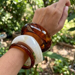 Op de afbeelding: Een set van drie gestapelde armbanden met een gladde, gepolijste afwerking. Eén armband is diep doorschijnend bruin, één is effen ondoorzichtig crèmewit en de derde is een dunnere, donkerbruine band met een glanzend, reflecterend oppervlak.