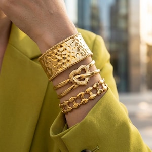 Può includere: Una collezione di braccialetti color oro. I braccialetti includono un'ampia manetta martellata, un design a forma di cuore e uno stile a catena. I braccialetti sono indossati su un polso e la persona indossa un blazer verde.