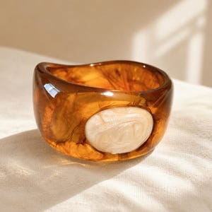 Può includere: Un bracciale rigido largo in resina traslucida color ambra, caratterizzato da un inserto ovale lucido color crema con motivi a vortice sulla parte anteriore. Il bracciale ha una forma organica liscia, arrotondata e leggermente irregolare.
