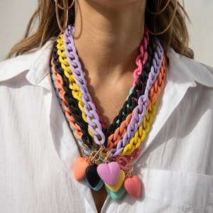 Può includere: Una collezione di collane a catena colorate in varie tonalità, tra cui nero, giallo, viola e rosa. Ogni collana è adornata da ciondoli a forma di cuore in colori coordinati, creando un accessorio vivace e giocoso.