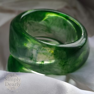 May include: A translucent green resin bangle bracelet. The wide band features swirling patterns of lighter green and white, creating a marbled effect. The bracelet is smooth and glossy, reflecting light.