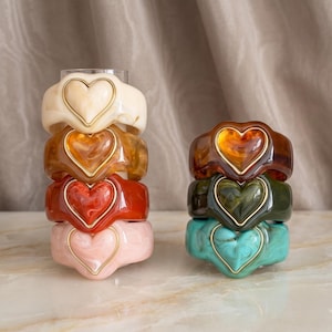May include: A collection of seven chunky resin bracelets stacked in two columns. Each bracelet features a prominent heart-shaped centre element with a gold-toned metal border. The colours include cream, amber, red, light pink, dark brown, olive, and teal.