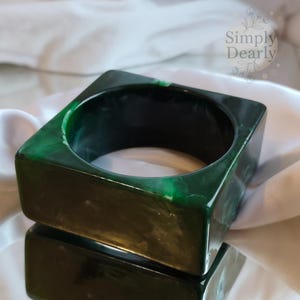 Può includere: Un braccialetto quadrato verde scuro con effetto marmorizzato. Il braccialetto ha una forma quadrata cava e si riflette sulla superficie sottostante. Il logo Simply Dearly è visibile sullo sfondo.
