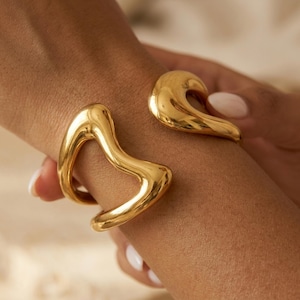Op de afbeelding: Een goudkleurige manchetarmband met een open ontwerp. De armband heeft een gladde, gepolijste afwerking en een sculpturale, organische vorm. De manchet wordt om een pols gedragen, wat de moderne uitstraling benadrukt. Het sieraad is een statement piece.