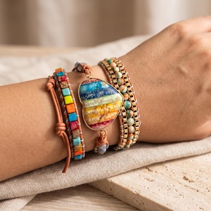 Può includere: Un bracciale avvolgente a più fili con un grande ciondolo a forma di cuore con bande orizzontali di pietra blu, verde, gialla, arancione e rossa. Il bracciale include perline quadrate e rotonde multicolori su cordini in pelle marrone.