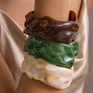 Può includere: Una pila di tre braccialetti rigidi spessi caratterizzati da una superficie lucida, irregolare e organica. Il braccialetto superiore è di un marrone traslucido intenso, quello centrale è verde bosco vivace e quello inferiore è bianco crema.