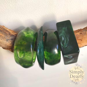 Può includere: Quattro braccialetti in resina verde con effetto marmorizzato, esposti su un pezzo di legno. I braccialetti hanno forme diverse, uno rettangolare e gli altri curvi. La resina è traslucida, permettendo alla luce di passare attraverso.