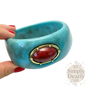 Può includere: Un bracciale a cerchio turchese con effetto marmorizzato. Presenta una grande gemma rossa ovale incastonata in una cornice di metallo dorato. Questo gioiello è un accessorio audace.