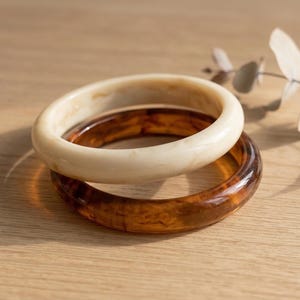 May include: Two stacked bangles. The top bangle is a creamy off-white color, while the bottom one is a translucent brown with a tortoiseshell pattern. The bangles are made of a smooth, glossy material and rest on a light-colored wooden surface.