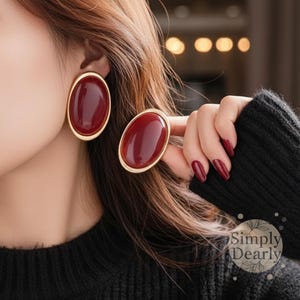 Puede incluir: Pendientes ovalados burdeos con borde dorado. Uno se lleva en la oreja y el otro lo sostiene una mano con esmalte de uñas burdeos a juego. Los pendientes tienen un acabado brillante y una estética vintage.
