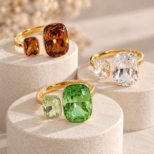 Può includere: Tre anelli aperti color oro, ciascuno con due pietre sfaccettate a taglio cuscino di dimensioni diverse. Un anello ha pietre color ambra, uno pietre bianche trasparenti e uno pietre verde brillante, montate su fasce di metallo lucido.